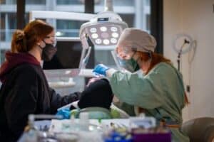 Surgeon in scrubs and cap performs a procedure on a patient, assisted by a masked colleague under a bright overhead surgical light in a clinical room.