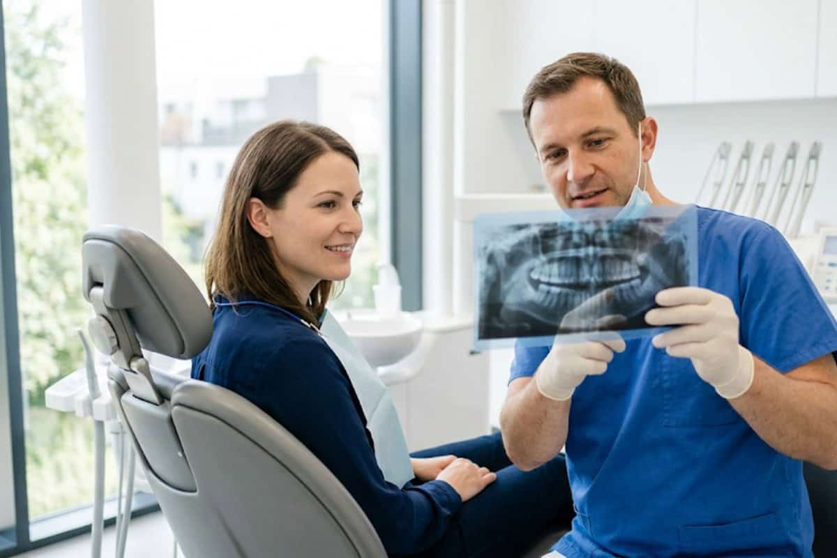 Dentist in blue scrubs shows a dental X-ray to a female patient sitting in a dental chair.